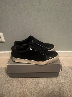 Lanvin skor - Snygga svarta sneakers med lågt skaft och klassisk design. Skorna har svarta skosnören, metallfärgade öljetter och en vit, något tjockare gummisula. Materialet ser ut att vara en mix av läder och mocka för en stilren look. Ganska slitna speciellt innersidan på hälen som man ser på dilferna. Storlek 10 eller 45