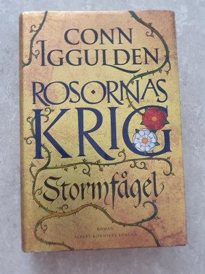 Bok - En historisk roman av Conn Iggulden med hård pärm och guldfärgat omslag. Boken handlar om maktkampen i England under 1400-talet och är första delen i serien Rosornas krig. Omslaget har dekorativa slingor och rosor i rött och vitt.