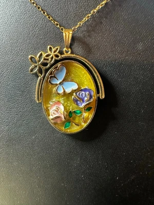 Guldigt halsband med fjäril och blommor - Unikt halsband med en oval, guldtonad berlock fylld med en blå fjäril och två blommor i rosa och lila, samt gröna blad. Berlocken har en glittrig gul bakgrund och dekoreras med små blommor upptill. Kedjan är tunn och matchar berlockens färg. Perfekt för dig som gillar färgglada detaljer.
