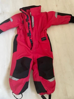 Röd overall från Polarn O. Pyret - Röd täckoverall från Polarn O. Pyret med svarta förstärkningar på knän, armbågar och bak. Reflexdetaljer på ärmar och ben för extra synlighet. Dragkedja framtill och resår i midjan samt benslut. Perfekt för kalla vinterdagar.