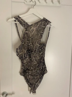 Bodysuit med ormmönster och omlott - Snygg bodysuit med trendigt ormmönster i beige och bruna toner. Omlott-design med breda axelband och draperad effekt framtill. Tillverkad i stretchigt, tunt material som sitter tajt mot kroppen. Perfekt för att skapa en cool och edgy look.