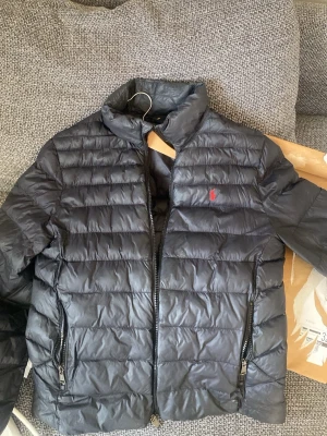 Svart pufferjacka från Polo Ralph Lauren - Svart pufferjacka från Polo Ralph Lauren med diskret röd logga broderad på bröstet. Jackan har ståkrage, två dragkedjefickor och quiltad design. Perfekt för kalla dagar och riktigt snygg till streetwear-stilen.