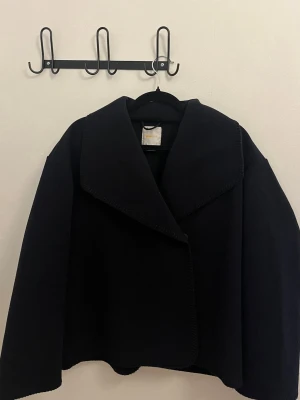 Marinblå kappa med bred krage - Stilren svart kappa med oversized passform och bred, markerad krage. Kappan har raka ärmar och en något kortare, boxig siluett. Perfekt för dig som gillar minimalistisk och modern stil. Materialet ser ut att vara tjockt och mjukt, troligen ullblandning.