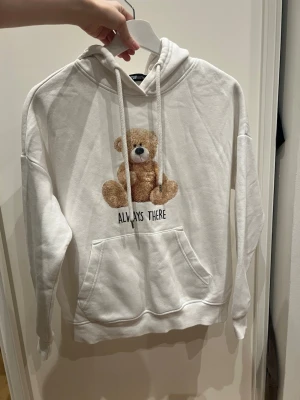 Vit hoodie med nalle och text - Mysig vit hoodie med huva och känguruficka framtill. På bröstet finns ett tryck av en söt brun nallebjörn och texten 'ALWAYS THERE' under. Hoodien har dragsko i huvan och är tillverkad i mjukt material, perfekt för en avslappnad och gullig stil.