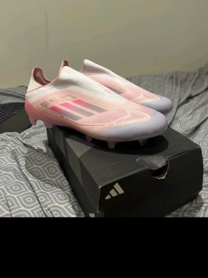 Adidas F50 Lamine Yamal elite fotbollskor - Snygga Adidas F50 fotbollsskor i en fräsch kombination av rosa och vitt med svarta detaljer. Skorna har en strömlinjeformad design och är tillverkade i syntetmaterial för lätthet och snabbhet på planen. Perfekta för dig som vill sticka ut och spela med stil.