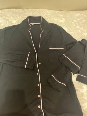 Svart pyjamas med rosa detaljer - Klassisk svart pyjamas med långärmad överdel och rosa kontrastkanter. Knäppning framtill med vita knappar och bröstficka med vit kant. Mjukt och bekvämt material, perfekt för mysiga kvällar hemma.