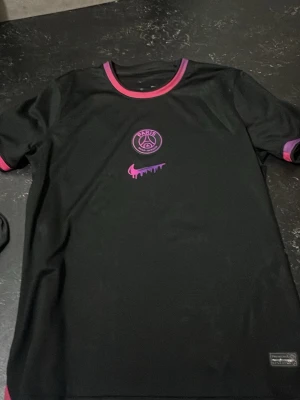 Svart PSG x Nike t-shirt - Säljer en svart PSG t-shirt från Nike med lila detaljer. Tröjan har PSG-logga och en unik lila Nike swoosh på bröstet. Rund halsringning och lila muddar vid ärmar och hals. Perfekt för dig som gillar fotboll och streetwear. Märket är gummi