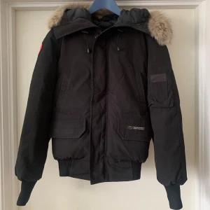Vintage Svart dunjacka från Canada Goose - Säljer en svart dunjacka från Canada Goose med icke avtagbar huva och äkta pälsdetalj. Jackan har ribbade muddar, flera fickor och den klassiska röda loggan på ärmen. Perfekt för kalla vinterdagar. Väldigt sparsamt använd och i väldigt bra skick. 