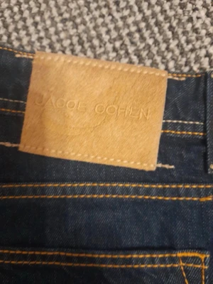 Jacob Cohen - Snygga mörkblå jeans från Jacob Cohen med klassisk femficksmodell och kontrastsömmar i gult. Jeansen har raka ben och en läderpatch med logga bak i midjan. Perfekta för dig som gillar stilrena och tidlösa jeans i denim.                  Riktigt bra skick!