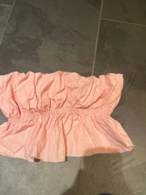 Ljusrosa bandeau topp - Söt ljusrosa bandeau topp med volang och resår i midjan. Toppen är axelbandslös och har en luftig, rynkad design som ger en avslappnad och trendig look. Perfekt för varma dagar och enkel att styla med jeans eller kjol.