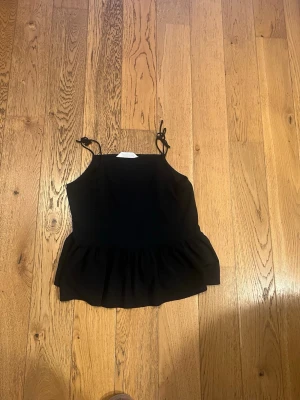 Svart linnetopp från H&M med volang - Snygg svart linnetopp från H&M med tunna axelband som knyts upptill och volang nedtill. Perfekt för en trendig och enkel look. Passar dig som gillar stilrena plagg med en twist.