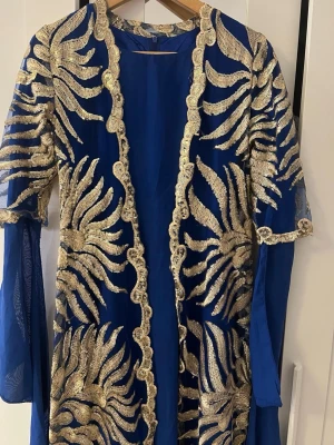 Kurdisk newroz kaftan klänning - Säljer en blå lång kappa med transparenta ärmar och detaljerade broderier i guld. Kappan har öppen front och ett unikt, festligt mönster som täcker hela plagget. Perfekt för dig som vill sticka ut med en elegant och färgstark look.