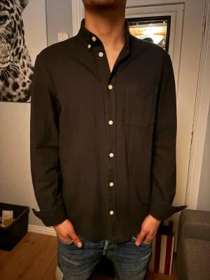 Svart skjorta med vita knappar - Stilren svart skjorta med button down-krage och vita kontrasterande knappar. Skjortan har en bröstficka och långa ärmar med manschetter. Perfekt till jeans för en avslappnad men snygg look.