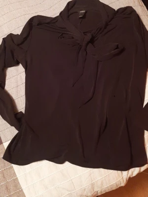 Svart blus med knyt från Lindex - Stilren svart blus från Lindex med knytband i halsen och långa ärmar. Blusen har en mjuk och lätt tygkänsla som ger ett snyggt fall. Perfekt för dig som gillar en klassisk och trendig look.