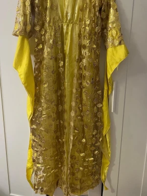 Kurdisk kafta newroz klänning  - Unik gul långklänning med glittriga paljettdetaljer och transparent tyg. Klänningen har vida ärmar och ett flowy fall som ger en riktigt statement look. Perfekt för dig som vill sticka ut och glänsa. Materialet känns lätt och skimrande.