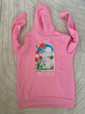 Rosa hoodie från Casablanca - Snygg rosa hoodie från Casablanca med färgglad tryckt design av rosor och landskap på både bröstet och ryggen. Hoodien har huva med dragsko och en stor magficka framtill. Perfekt för dig som vill sticka ut med en unik och trendig stil.Storlek M eller S