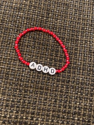 Rött pärlarmband med ADHD-bokstäver - Snyggt armband med små röda pärlor och vita bokstavspärlor som stavar ADHD. Armbandet är elastiskt och passar de flesta handleder. Perfekt accessoar för dig som vill uttrycka din personlighet med stil.