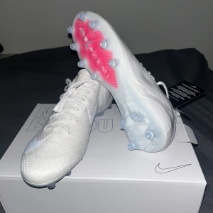 Nike by you  vita fotbollsskor Air Zoom - Säljer ett par grymt snygga vita Nike Air Zoom fotbollsskor med snörning och platt sula. Skorna har en modern design med transparenta dobbar och en hint av rosa under sulan. Perfekta för dig som vill sticka ut på planen med stil och komfort.