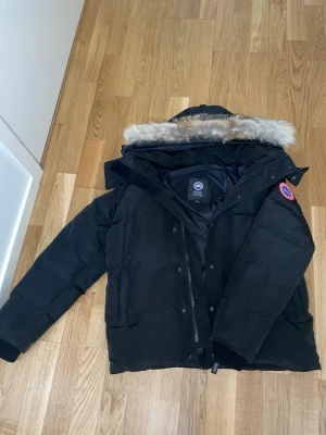 Svart Canada Goose jacka  - Säljer en svart Canada Goose jacka med avtagbar huva och pälsdetalj. Jackan har en klassisk passform, tydlig logga på ärmen och dragkedja med knappar framtill. Perfekt för kalla vinterdagar och riktigt varm.
