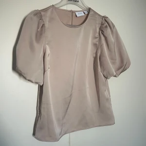 Beige puffärm blus från VILA - Stilren beige blus från VILA med puffiga korta ärmar och rund halsringning. Tillverkad i ett glansigt, mjukt satin material som ger en elegant känsla. Perfekt för dig som vill ha en enkel men trendig look. Oanvänd.