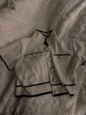 Vit croppad cardigan med pärlknappar sett - Snygg vit croppad cardigan med svarta detaljer och dekorativa pärlknappar framtill. Tröjan har korta ärmar, två fejkfickor och ribbade kanter. Perfekt för en trendig och chic look. Kjolen medföljer 