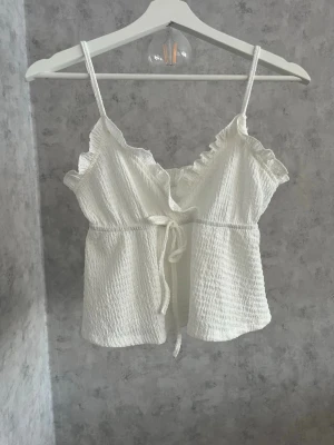 Vit linnetopp med volang och knyt - Söt vit linnetopp med smala axelband, volangdetaljer vid urringningen och knyt i midjan🪽🪽
