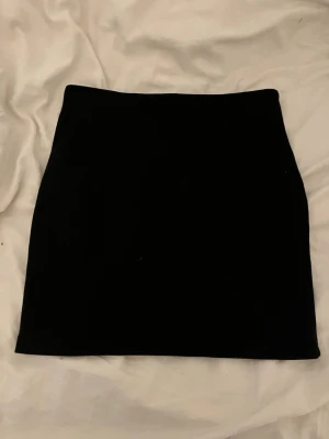 Svart kortkjol i enkel design - En stilren svart kortkjol med rak passform. Kjolen har en minimalistisk look utan synliga detaljer och passar perfekt till många olika outfits. Tillverkad i ett mjukt material som känns bekvämt mot huden.