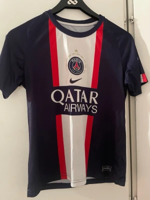 PSG matchtröja Mbappé Nike - Snygg PSG matchtröja i mörkblått med röda och vita detaljer. Framsidan har Paris Saint-Germain-logga, Qatar Airways-tryck och Nike swoosh. Baksidan har Mbappé och nummer 7. Tillverkad i lätt och ventilerande material, perfekt för fotboll eller träning. Nypris 399kr, säljs för 60kr