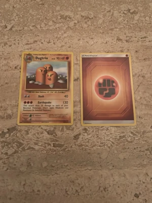 Två Pokémonkort: Dugtrio & Fighting Energy - Säljer två Pokémonkort: ett Dugtrio-kort med 90 HP och attacker samt ett Fighting Energy-kort. Båda har klassisk design med gul ram och färgstarka motiv. Perfekt till samlingen eller för att spela Pokémon TCG med vänner.