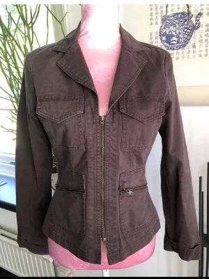 Brun/grön  utilityjacka med dragkedja - Snygg brun/grön  utilityjacka med markerad midja och fyra fickor framtill. Jackan har dragkedja, klassisk krage och långa ärmar. Tillverkad i bomullsmaterial som ger en avslappnad men ändå strukturerad look.