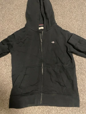 Svart hoodie med dragkedja från Gant - Svart hoodie från Gant med dragkedja framtill, huva och två stora fickor. Liten broderad Gant-logga på bröstet. Klassisk och stilren design med ribbade muddar och mjukt material, perfekt för en avslappnad look. Väldigt bra skick och den passar folk mellan 156-162cm