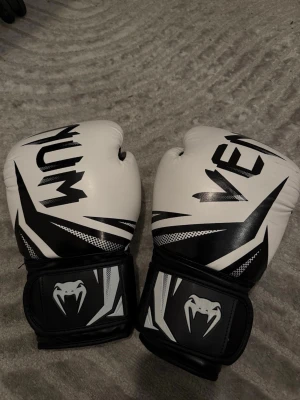 Venum boxningshandskar svart/vit - Säljer ett par boxningshandskar från Venum i svart och vitt med coolt mönster och logga på handleden. Handskarna har kardborreknäppning och är tillverkade i syntetmaterial. Perfekta för träning inom boxning eller kampsport.har använt dem bara 2 gånger