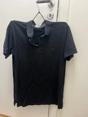 Svart pikétröja från Ralph Lauren - Klassisk svart pikétröja från Ralph Lauren med korta ärmar och krage. Tröjan har knappslå framtill och den ikoniska broderade loggan på bröstet. Tillverkad i mjuk bomull som ger en avslappnad och stilren look.