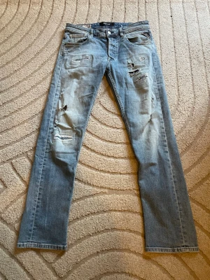 Ljusblå slitna jeans från Replay - Säljer ett par ljusblå jeans från Replay med slitningar och trasiga detaljer på både fram- och baksida. Jeansen har klassisk femficksdesign, raka ben och knappgylf. Perfekta för en avslappnad och trendig look.