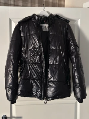 Svart dunjacka från Moncler - Snygg svart dunjacka från Moncler med glansig finish och quiltat mönster. Jackan har huva, dragkedja framtill och två sidofickor med dragkedja. Perfekt för kalla vinterdagar och riktigt trendig look.