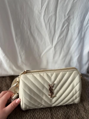 Vit handväska inte äkta  - Inte äkta.   Vit väska med guld detaljer & dragkedja två små innerfickor & långt axelband 