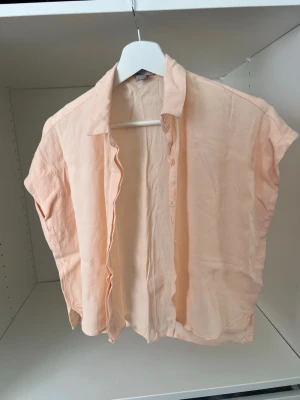 Ljus persikofärgad blus med krage - Säljer en ljus persikofärgad blus med korta ärmar och klassisk krage. Blusen har knappar framtill och är tillverkad i ett tunt, mjukt tyg som ger en avslappnad känsla. Perfekt för en enkel och stilren look.
