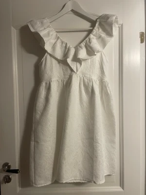 Vit kortklänning med volang - Supersöt vit kortklänning med bred volang runt axlarna och v-ringning. Klänningen har ett diskret mönster i tyget och är ärmlös. Perfekt för student och sommarens alla tillfällen!