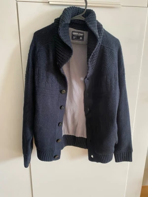 Mörkblå stickad kofta Green Coast - Mysig mörkblå stickad kofta från Green Coast med grovstickad krage och knappar framtill. Koftan har ribbade muddar vid ärmslut och nederkant samt diskret mönsterstickning upptill. Perfekt för lager på lager och chill stil.