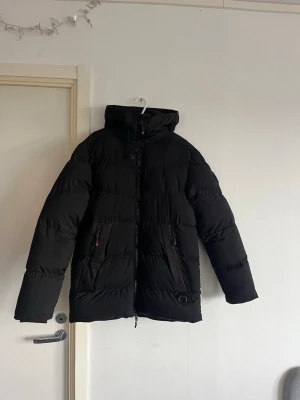 Svart pufferjacka med huva - Säljer en svart pufferjacka med hög krage och huva. Jackan har två dragkedjeförsedda fickor framtill och justerbar dragsko vid huvan. Perfekt för kalla vinterdagar och riktigt skön att bära. Klassisk och stilren design som passar till det mesta.