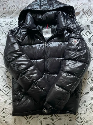 Svart pufferjacka från Moncler - Snygg svart pufferjacka från Moncler med glansig finish, huva och dragkedja framtill. Jackan har en patch med Moncler-logga på ärmen och två sidofickor med dragkedja. Perfekt för kalla vinterdagar och riktigt trendig look.