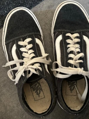 Svarta Vans Old Skool sneakers - Klassiska Vans Old Skool sneakers i svart mocka och canvas med vita detaljer och den ikoniska sidoranden. Skorna har vita snören och en vit gummisula. 