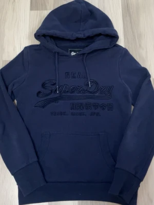 Superdry hoodie LIMITED edition  - Snygg mörkblå hoodie från Superdry med broderad logga och japanska tecken på bröstet. Klassisk känguruficka framtill, ribbade muddar och justerbar huva med dragsko. Perfekt för en avslappnad och trendig stil.