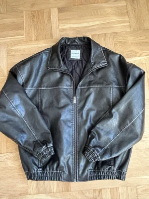 Mörkbrun skinnjacka från Bershka - Snygg mörkbrun skinnjacka från Bershka med dragkedja framtill, klassisk krage och resår vid ärmslut och midja. Jackan har en stilren look och är fodrad för extra komfort. Perfekt för dig som vill ha en trendig och tidlös jacka i garderoben.