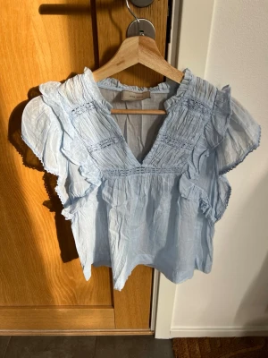 Superfin Ljusblå volangblus från Neo Noir, stl 36 - Supersöt ljusblå blus från Neo Noir med volangdetaljer och v-ringning. Blusen har korta ärmar med rynkade partier och är gjord i ett tunt, luftigt tyg som känns perfekt för sommaren. Romantisk look med fina sömmar och lätt struktur. Aldrig använd, helt ny, perfekt till skolavslutningen🌸☀️! 