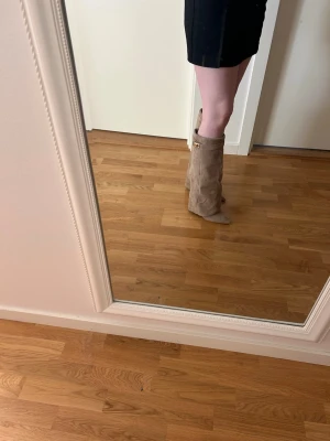 Beige mockastövlar med slouchy skaft - Säljer ett par snygga beige stövlar i mocka med slouchy skaft och spetsig tå. Stövlarna har en dekorativ gulddetalj upptill och passar perfekt till kjol eller klänning. Klackhöjden är 9cm och materialet känns mjukt och lyxigt. Inte använda än enda gång.