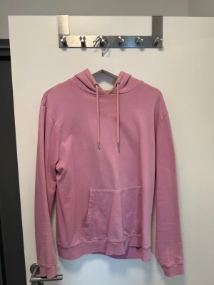 Rosa hoodie från ASOS med huva - Säljer en rosa hoodie från ASOS i storlek M. Hoodien har en klassisk huva med dragsko, stor magficka och ribbade muddar vid ärmslut och nederkant. Tillverkad i mjukt bomullsmaterial som är skönt mot huden. Perfekt för en chill och avslappnad stil.