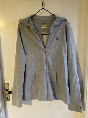 Grå zip hoodie från U.S. Polo Assn. - Snygg grå hoodie med dragkedja från U.S. Polo Assn. Klassisk design med huva, två fickor framtill och liten broderad logga på bröstet. Perfekt till ett par jeans. Finns en liten fläck på vänster ärm men inget man tänker på (se sista bilden). Storlek  14-15 och sitter som xs. Nypris 649kr. 💕