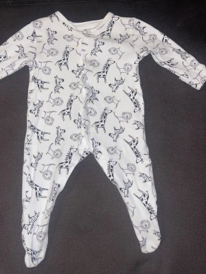 Vit sparkdräkt med djurmönster Primark - Supermjuk vit sparkdräkt från Primark Baby med tryck av zebror, lejon och giraffer i svart och gult. Långa ärmar och ben med inbyggda fötter samt tryckknappar framtill för enkel påklädning. Perfekt för mysiga stunder hemma.