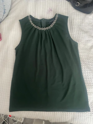 Mörkgrön ärmlös topp med pärlor - Snygg mörkgrön ärmlös topp från Stockh lm med dekorativa pärlor runt halsringningen. Toppen har en rak passform och knäppning i nacken. Perfekt för dig som vill ha något stilrent men ändå med en twist.